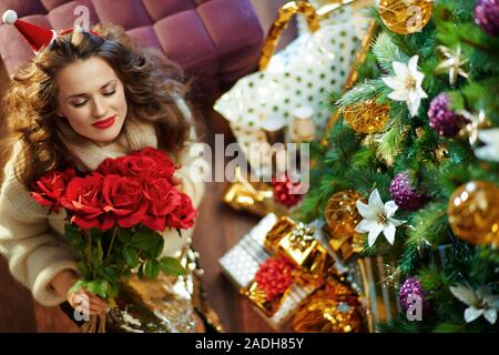 Obere Ansicht von lächelnden trendy mittleren Alter Frau mit langen brünetten Haar in Gold Pailletten Rock und weißen Pullover unter dem geschmückten Weihnachtsbaum in der Nähe von prese Stockfoto