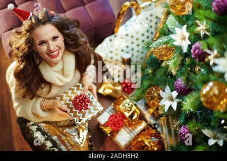 Obere Ansicht von lächelnden stilvolle Frau mit langen brünetten Haar in Gold Pailletten Rock und weißen Pullover unter dem geschmückten Weihnachtsbaum in der Nähe des heutigen Boxen h Stockfoto