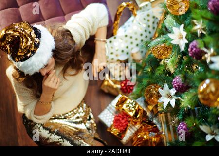 Obere Ansicht von besorgt trendy Hausfrau mittleren Alters mit langen brünetten Haar in Gold Pailletten Rock und weißen Pullover unter dem geschmückten Weihnachtsbaum in der Nähe von p Stockfoto