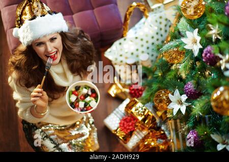 Obere Ansicht von Happy trendy Hausfrau mit langen brünetten Haar in Gold Pailletten Rock und weißen Pullover unter dem geschmückten Weihnachtsbaum in der Nähe des heutigen Boxen Stockfoto