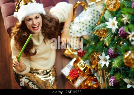 Obere Ansicht von lächelnden trendy Frau mit langen brünetten Haar in Gold Pailletten Rock und weißen Pullover mit sellerie unter dem geschmückten Weihnachtsbaum in der Nähe von Pres Stockfoto