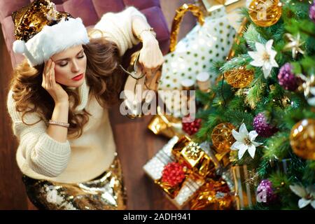 Obere Ansicht der Müde trendy 40 Jahre alte Frau mit langen brünetten Haar in Gold Pailletten Rock und weißen Pullover unter dem geschmückten Weihnachtsbaum in der Nähe von presen Stockfoto