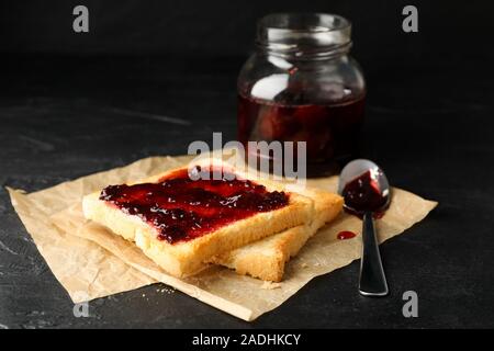 Toast und Marmelade, Glas Glas mit Löffel und Backpapier auf schwarzem Hintergrund, in der Nähe Stockfoto