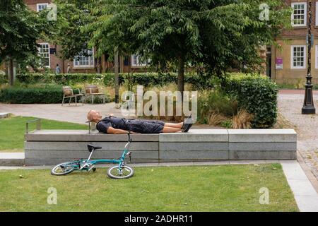 Junge Mann in Shorts Schlafen auf dem Rücken auf einem konkreten Sitz, mit seinem Fahrrad auf dem Boden. England, Großbritannien Stockfoto