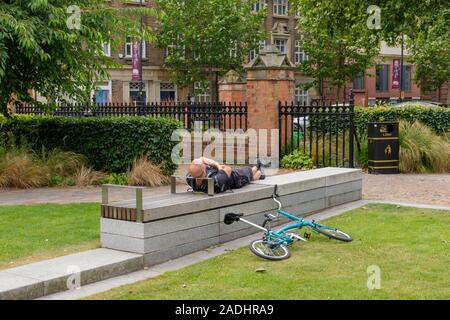 Junge Mann in Shorts Schlafen auf dem Rücken auf einem konkreten Sitz, mit seinem Fahrrad auf dem Boden. England, Großbritannien Stockfoto