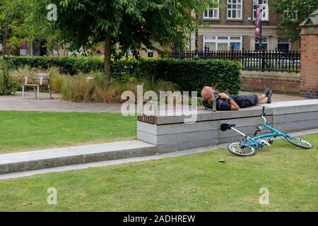 Junge Mann in Shorts Schlafen auf dem Rücken auf einem konkreten Sitz, mit seinem Fahrrad auf dem Boden. England, Großbritannien Stockfoto