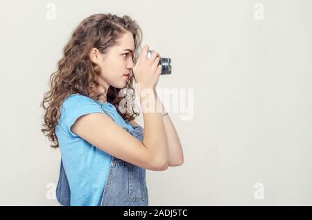 Curly brunette Mädchen in Jeans Overalls hält ein Vintage film Kamera in den Händen. Nimmt ein Bild. Von der Seite. Stockfoto