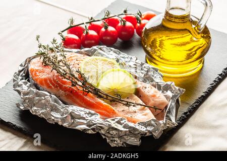 Fisch in Folie mit Thymian und Limetten auf schwarzem Schiefer platziert Stockfoto