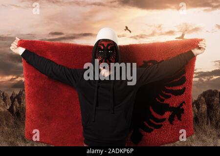 Junger Mann mit paintad albanische Flagge auf dem Gesicht stand vor der felsigen Landschaft und fliegen die flauschigen albanische Flagge Stockfoto