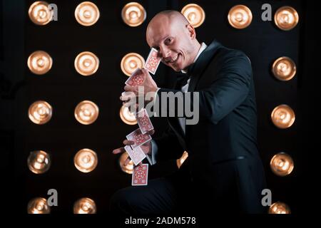 Lächelnd während des Prozesses. Magier im schwarzen Anzug und mit Spielkarten im Zimmer stehend mit spezieller Beleuchtung auf der Bühne Stockfoto