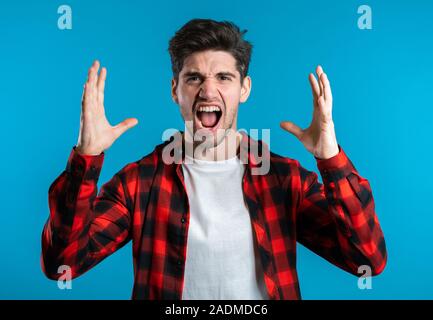 Junge europäische betonte Mann im roten Hemd Schreien über blauen Hintergrund isoliert. Gestresst und deprimiert Kerl laut schreiend auf die Kamera. Stockfoto