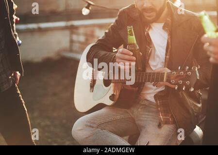 Bärtiger Kerl sitzt mit akustischer Gitarre auf dem Dach in Nizza Herbst Tag und halten nur neue Bier geöffnet Stockfoto