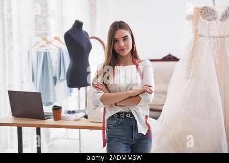 Mit verschränkten Armen und einem Tisch mit Laptop. Weibliche Mode Designer arbeitet auf der neuen Kleidung in der Werkstatt Stockfoto