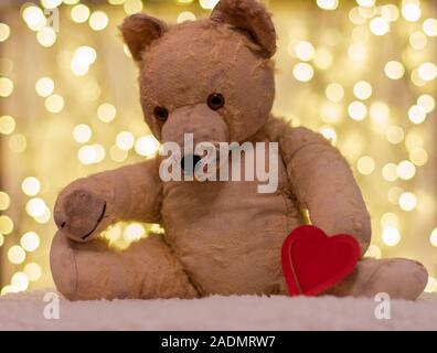 Sehr alte Soft Toy. Zwischen 40 bis 50 Jahre alten Teddybär mit rotem Herz aus Papier. Auf dem Hintergrund sind Weihnachtsbeleuchtung. Sehnsucht, Liebe. Stockfoto