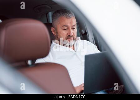 Sitzen in der luxuriösen Fahrzeug. Arbeiten an einem Auto mit silbernen Laptop. Senior Geschäftsmann Stockfoto