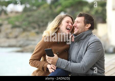 Glückliches Paar von Erwachsenen lacht laut über Handy Content sitzend auf dem Strand im Winter Stockfoto