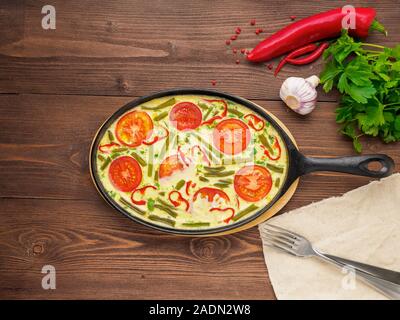 Pfanne mit Frittata in der Mitte auf einem braunen Holztisch, Gabeln, Petersilie, Pfeffer, Tomaten, Ansicht von oben Stockfoto