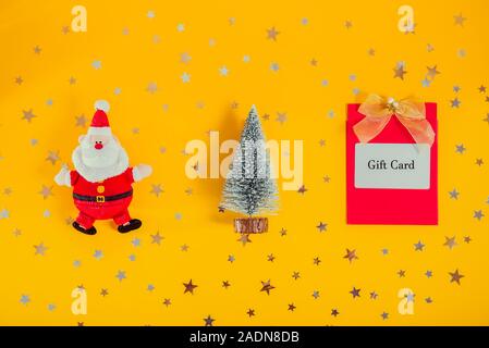 Ansicht von oben festliche Komposition mit Weihnachtsmann, Weihnachtsbaum und Geschenkkarte auf gelben Hintergrund mit funkeln Sterne. Kreative geschenk idee. Neue Stockfoto
