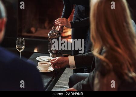 Frau Kellner strömenden heißen Tee in die weiße Schale. Freunde sitzt in einem Restaurant mit schönem Kamin in Es Stockfoto