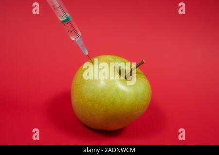 Gift dringt in den grünen apfel ein. Spritze mit Flüssigkeit in Obst. Foto des Konzepts "GMO". Vergiftete Lebensmittel auf rotem Hintergrund. Nahaufnahme. Stockfoto