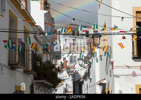 Eine Straße in Sedella mit Bunting für Saint Anthony feiern im Januar eingerichtet. Ein Regenbogen erscheint am Himmel. Region Axarquia, Andalusien, Spanien. Stockfoto