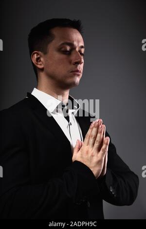 Ernsthafte denken Mann, der betet und gestering die Hände im schwarzen Anzug auf der grauen Schatten studio Hintergrund. Closeup Portrait. Stockfoto