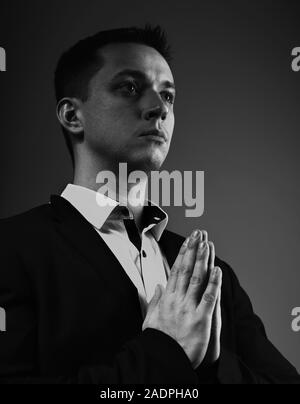 Ernsthafte denken Mann, der betet und gestering die Hände im schwarzen Anzug auf der grauen Schatten studio Hintergrund. Closeup Portrait. Schwarz und Weiß Stockfoto