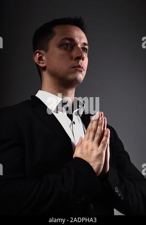 Ernsthafte denken Mann, der betet und gestering die Hände im schwarzen Anzug auf der grauen Schatten studio Hintergrund. Closeup Portrait. Stockfoto