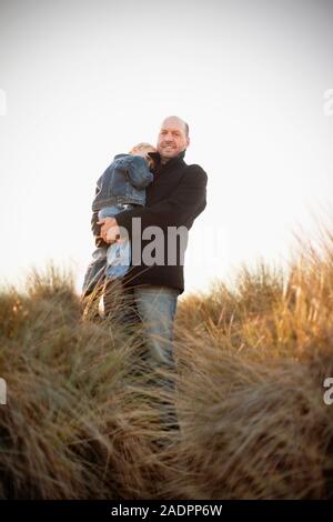Mann lächelt, als er seinen kleinen Sohn, Knuddel in seine Schulter, als sie für ein Portrait auf einem grasbewachsenen Düne Pose hält. Stockfoto