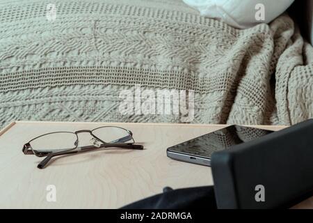 Gläser, Fall- und Smartphone auf dem Nachttisch im Schlafzimmer. Brillen Lagerung und Pflege. Selektiver Fokus Stockfoto