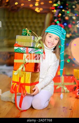 Ein lustiges Mädchen in a Gnome's Kappe sitzt auf einer Hälfte mit vielen Geschenken in den Händen vor dem Hintergrund der hellen Holiday Lights. Weihnachten und neues Jahr. Stockfoto