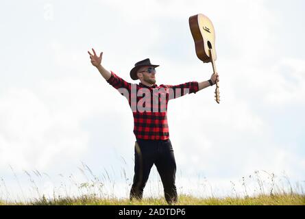 Inspiriert land Musiker. Landhausstil. Sommer Urlaub. Wandern Song. United mit der Natur. Stattlicher Mann mit Gitarre. Country Music Konzept. Gitarrist Land Sänger stand im Feld Himmel Hintergrund. Stockfoto