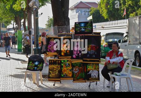 Bangkok, Thailand - 2. Dezember 2019: Eine weibliche Street Hersteller sitzen auf Stuhl vor Wat Pho, einem berühmten Tempel, Verkauf von Andenken Gemälde Stockfoto