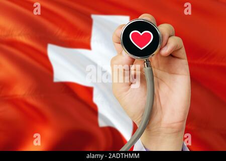 Arzt holding Stethoskop mit roten Liebe Herz. Nationale Schweiz Flagge Hintergrund. Healthcare System Konzept, medizinische Thema. Stockfoto