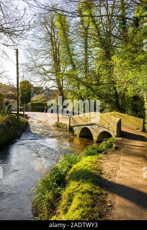 Die alte Stein Fuß-Brücke überspannt den langsam fließenden ByBrook stream, das Bestandteil eines Ford für vehicular Kreuzungen im ruhigen Dorf Lacock i Stockfoto