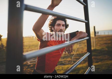 Jungen athletischen Mann eine Pause vom Training auf den vertikalen Balken im Fitnessbereich im Freien Park am Morgen