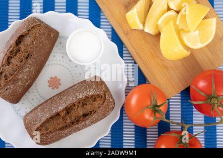 Schwarze Brötchen auf einem weissen Teller, Salz, Zitronen und Tomaten. Snacks, leichte Mahlzeiten, nosh. Stockfoto
