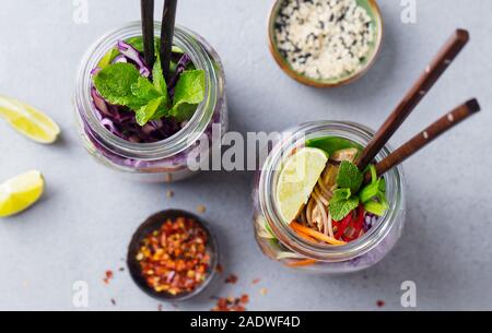 Soba Nudeln Salat, Suppe mit Gemüse, Tofu und Huhn in Gläsern. Grauer Hintergrund. Ansicht von oben. Stockfoto