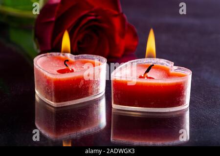 Nahaufnahme von zwei brennende Herz Valentinstag Kerzen mit rote Rose im Hintergrund auf einem dunklen Tabelle Stockfoto