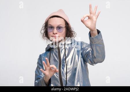 Froh, attraktive Frau in Gläsern und rosa Hut zeigt ok Zeichen als Zustimmung ausdrückt, Gesten im Studio hat fröhlichen Ausdruck. Stockfoto