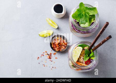 Soba Nudeln Salat, Suppe mit Gemüse, Tofu und Huhn in Gläsern. Grauer Hintergrund. Kopieren Sie Platz. Ansicht von oben Stockfoto