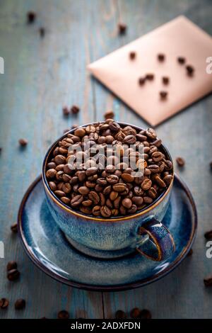 Flach Cappuccino Tasse mit Kaffee Bohnen auf den Tisch verstreut, blau Kaffeetasse obere Ansicht Nahaufnahme auf Holz- Hintergrund in der Sonne Licht. Cafe und Bar, Barist Stockfoto