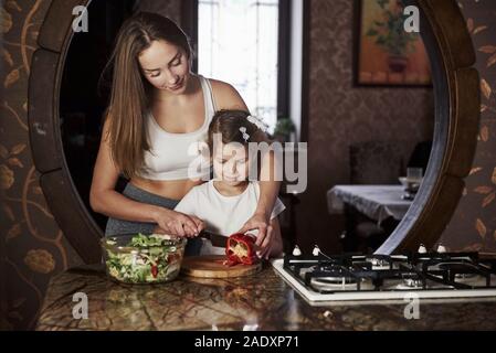 Versucht, eine Schicht Gemüse. Hübsche junge Frau, die in der modernen Küche in der Nähe von Gas- Herd und Tochter beibringen, wie man Essen zubereiten Stockfoto