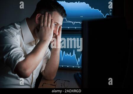 Belastende Situation. Bärtiger Mann im weißen Hemd arbeitet im Büro mit mehreren Bildschirmen in Index charts Stockfoto