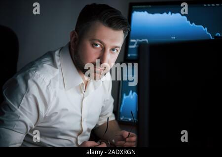 Der Blick in die Kamera. Bärtiger Mann im weißen Hemd arbeitet im Büro mit mehreren Bildschirmen in Index charts Stockfoto