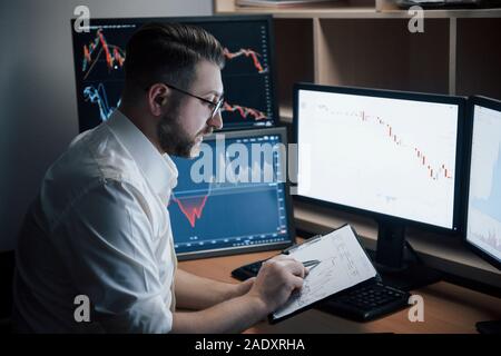 Mit Notepad. Bärtiger Mann im weißen Hemd arbeitet im Büro mit mehreren Bildschirmen in Index charts Stockfoto