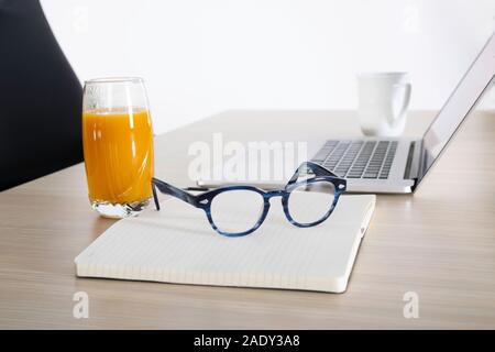 Laptop auf einem hölzernen Schreibtisch mit offenen Notebook, Lesebrille, Glas Saft und eine Tasse Kaffee. Stockfoto