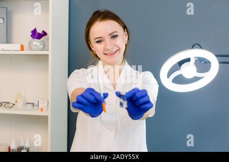 Junge Frau in weißem Kleid und blaue Handschuhe hält Spritze in Arztpraxis, bereit für die Injektion. Stockfoto