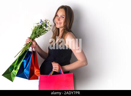 Eine schwangere Mädchen steht gegen die Wand mit Blumen und Taschen aus dem Store. Freudige schwangere Frau mit Einkaufstüten holding Blumenstrauß Stockfoto