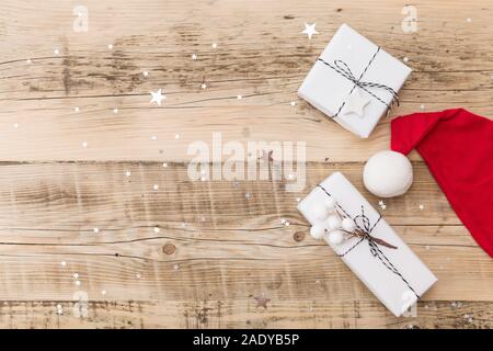 Draufsicht auf schöne Weihnachten Geschenke verpackt in Weiß Geschenkpapier, Christbaumschmuck in Santa's Hut auf Holz- Hintergrund mit funkelnden Sternen. Neue Stockfoto